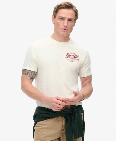 Cream-colored patterned T-shirt featuring a vintage Superdry logo in red, made from soft cotton, with short sleeves and a relaxed fit, paired with khaki pants and a dark green sweater tied at the waist.