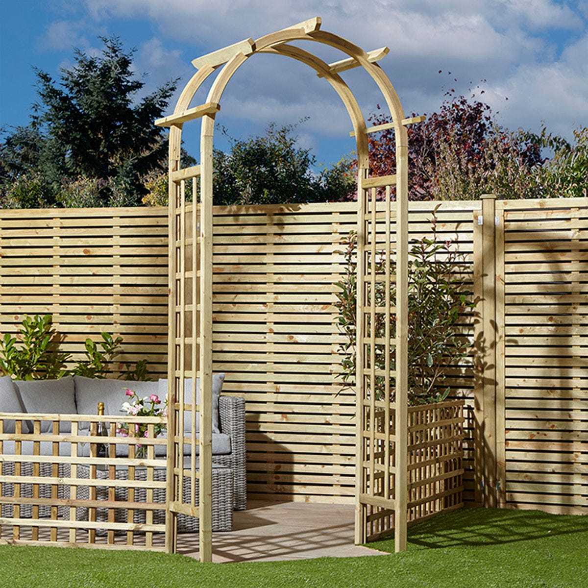 Natural pressure-treated timber arch with a curved top and trellis sides, set against a wooden fence, surrounded by greenery and outdoor furniture.