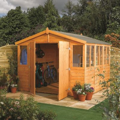 A brown wooden workshop measuring 9ft x 12ft with double doors, large windows, and a sloped roof, surrounded by greenery and potted plants.