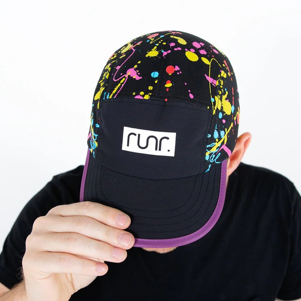 Black cap with vibrant splatter paint design in pink, blue, and yellow, featuring a white "runr." logo on the front and a purple visor, made from technical running material.
