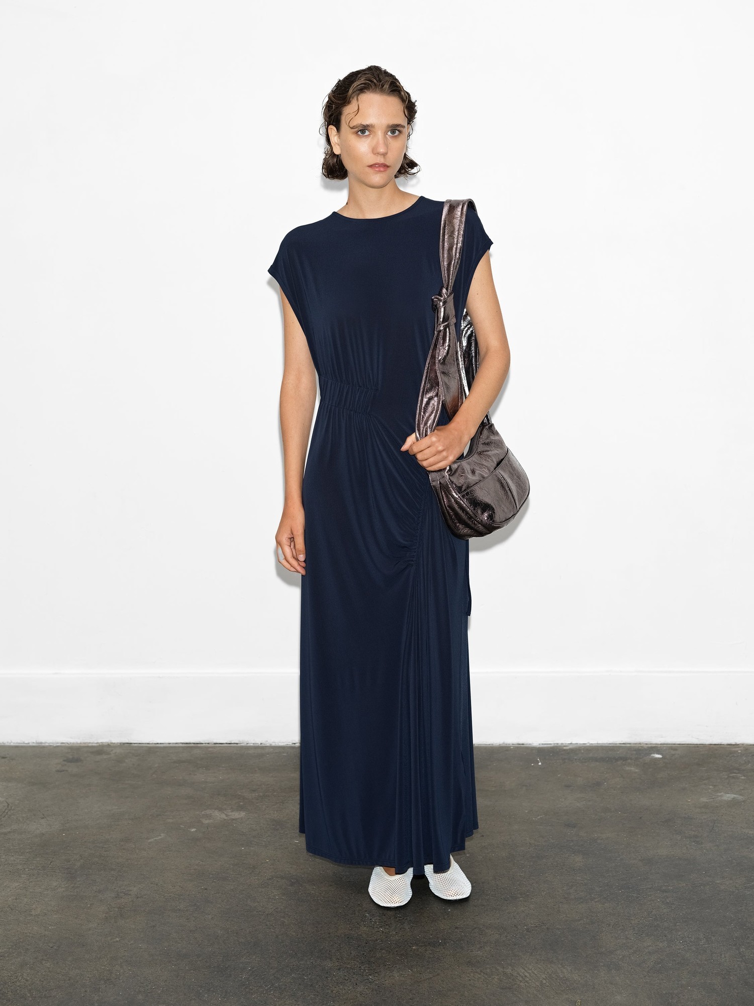 Navy gathered maxi dress made of soft jersey fabric, featuring short sleeves, a cinched waist, and a flowing skirt, paired with a metallic shoulder bag and white shoes.