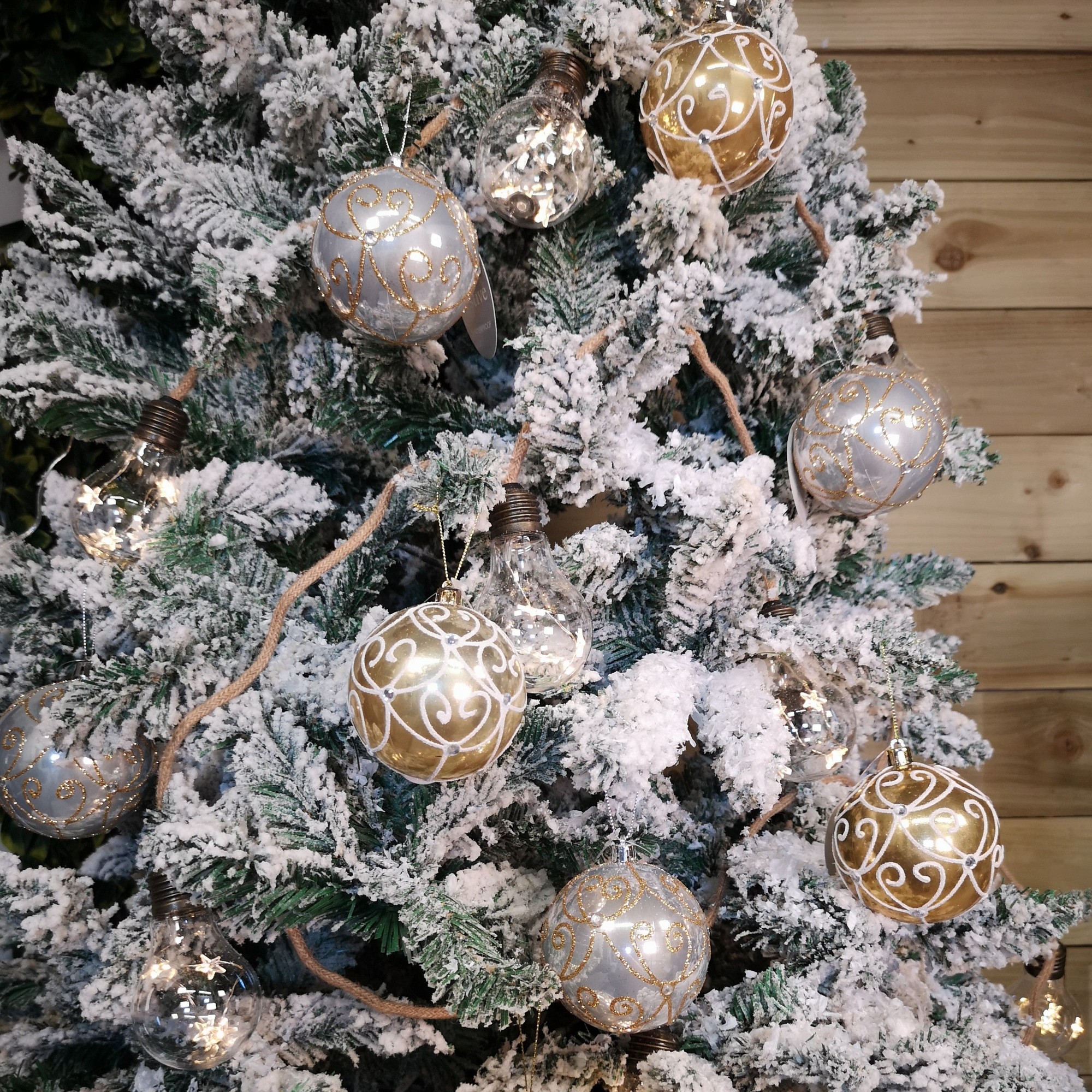 Box of 12 shatterproof Christmas baubles in assorted pearl gold and silver, featuring intricate designs, hanging on a snowy green tree.