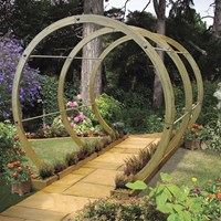 A brown wooden garden arch with curved beams, set on a stone pathway surrounded by colorful flowers and greenery, creating an inviting outdoor space.