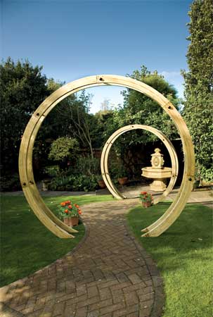 Two large, circular wooden arches made from light-colored timber stand in a garden, surrounded by greenery and a stone fountain in the background.
