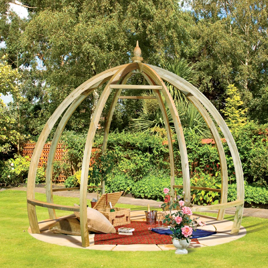 A wooden Grange Apollo Pergola with a dome shape, featuring natural timber, open sides, and a decorative finial, set on a grassy area surrounded by plants and flowers.