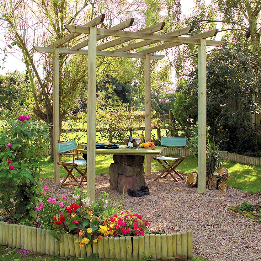 Brown wooden Grange Traditional Pergola with a slatted roof, surrounded by colorful flowers and greenery, featuring two chairs and a table set in a gravel area.