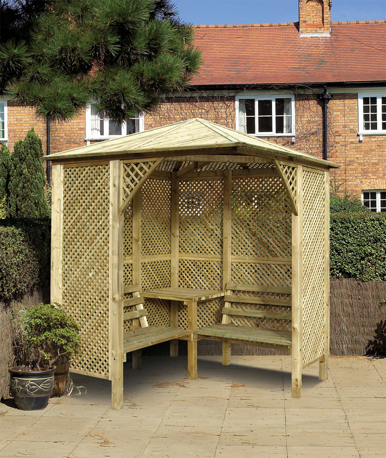 Wooden corner arbour seat with a table, featuring a decorative trellis design, set in a garden with brick buildings and greenery in the background.