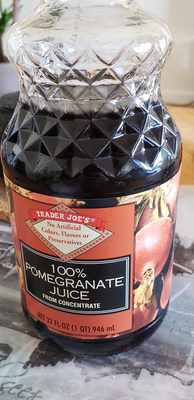 Clear glass bottle with a diamond-patterned lid, filled with dark red 100% pomegranate juice, featuring a label with orange and black design and text stating "No Artificial Colors, Flavors or Preservatives."