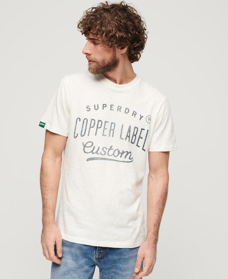 Light beige Superdry T-shirt featuring "Copper Label" and "Custom" printed in dark gray, with a green logo tag on the sleeve and a relaxed fit.