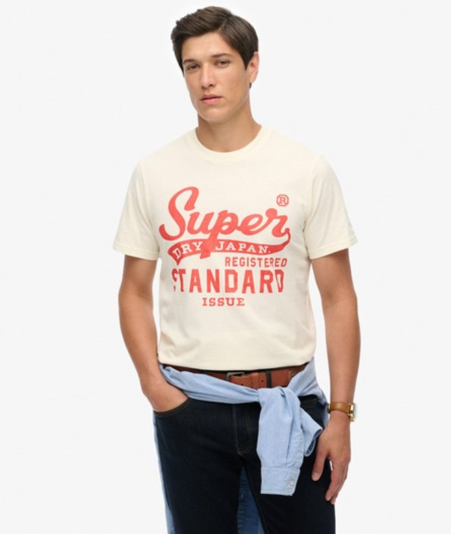 Cream-colored T-shirt featuring bold red "Superdry" script and "Standard Issue" text, made of soft cotton, worn with a light blue sweater tied around the waist.