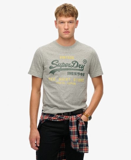 Grey cotton t-shirt featuring a large vintage Superdry logo in green and yellow, with a relaxed fit and short sleeves, styled with a plaid shirt tied around the waist.