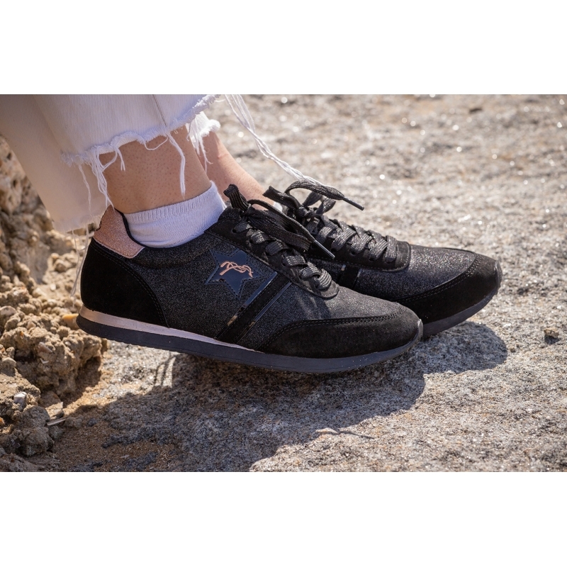 Black glitter trainers with a suede finish, featuring a pink logo and a sleek design, worn with white distressed jeans on a rocky surface.