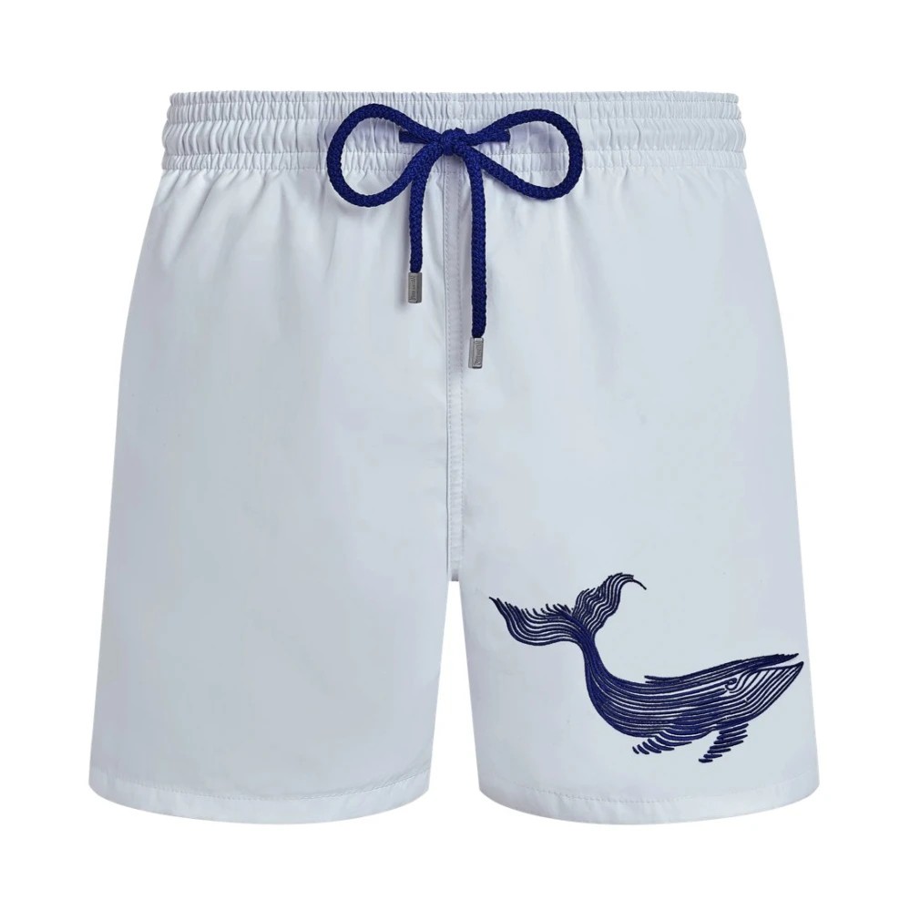 White swim shorts featuring an embroidered blue whale design on the left side, with an elastic waistband and a contrasting blue drawstring.