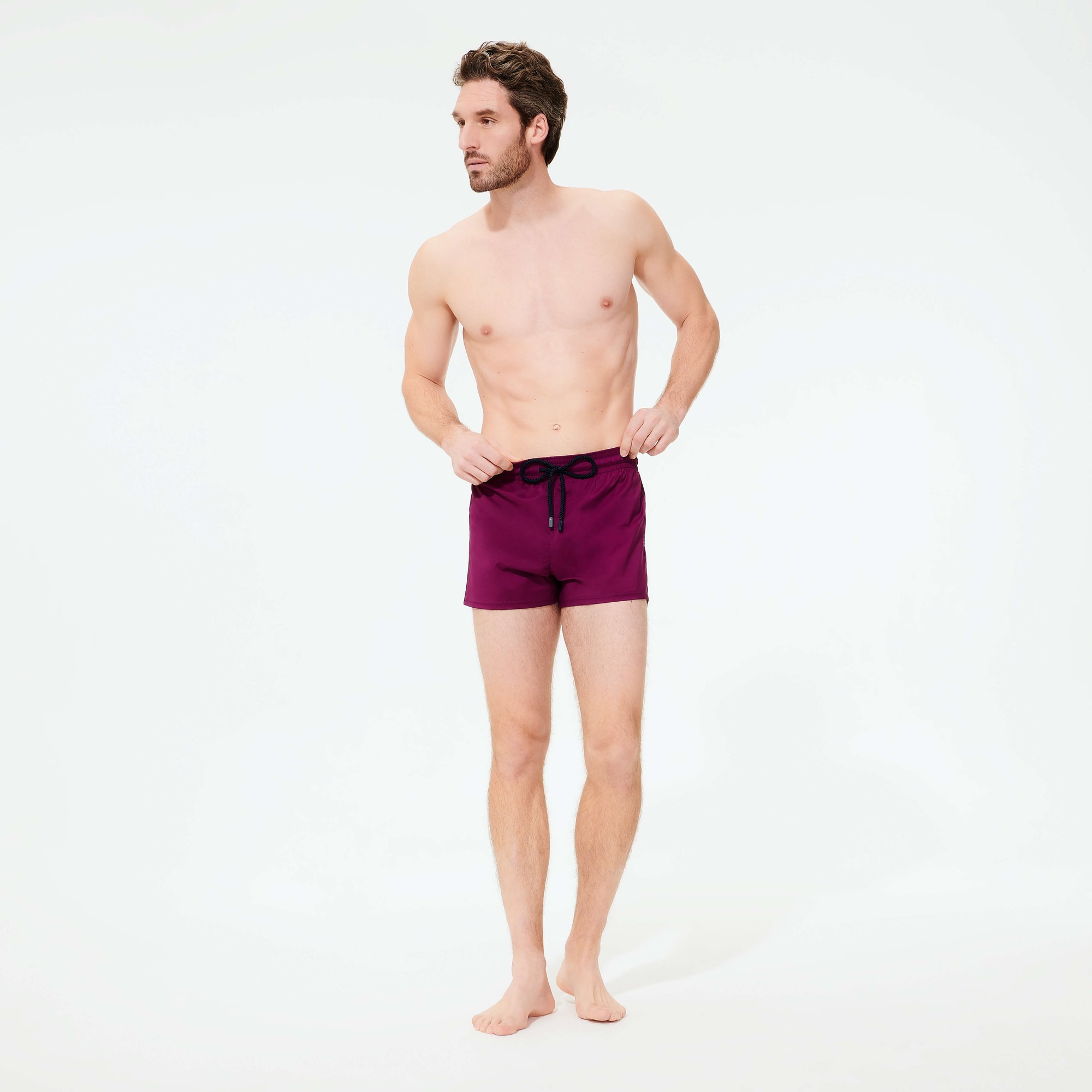 Men's fitted swim shorts in solid purple, featuring an elastic waistband with a drawstring, made from stretch material, displayed on a model standing barefoot.