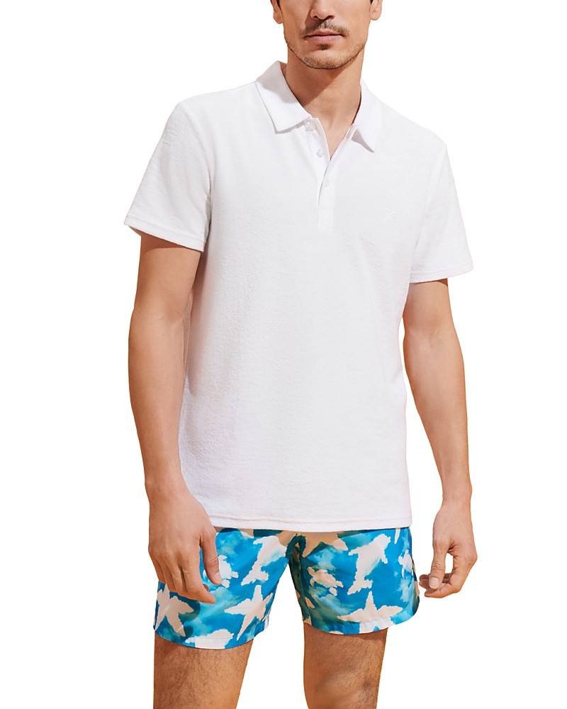 White terry polo shirt by Vilebrequin, featuring a classic collar and short sleeves, paired with colorful blue swim shorts, model stands confidently.