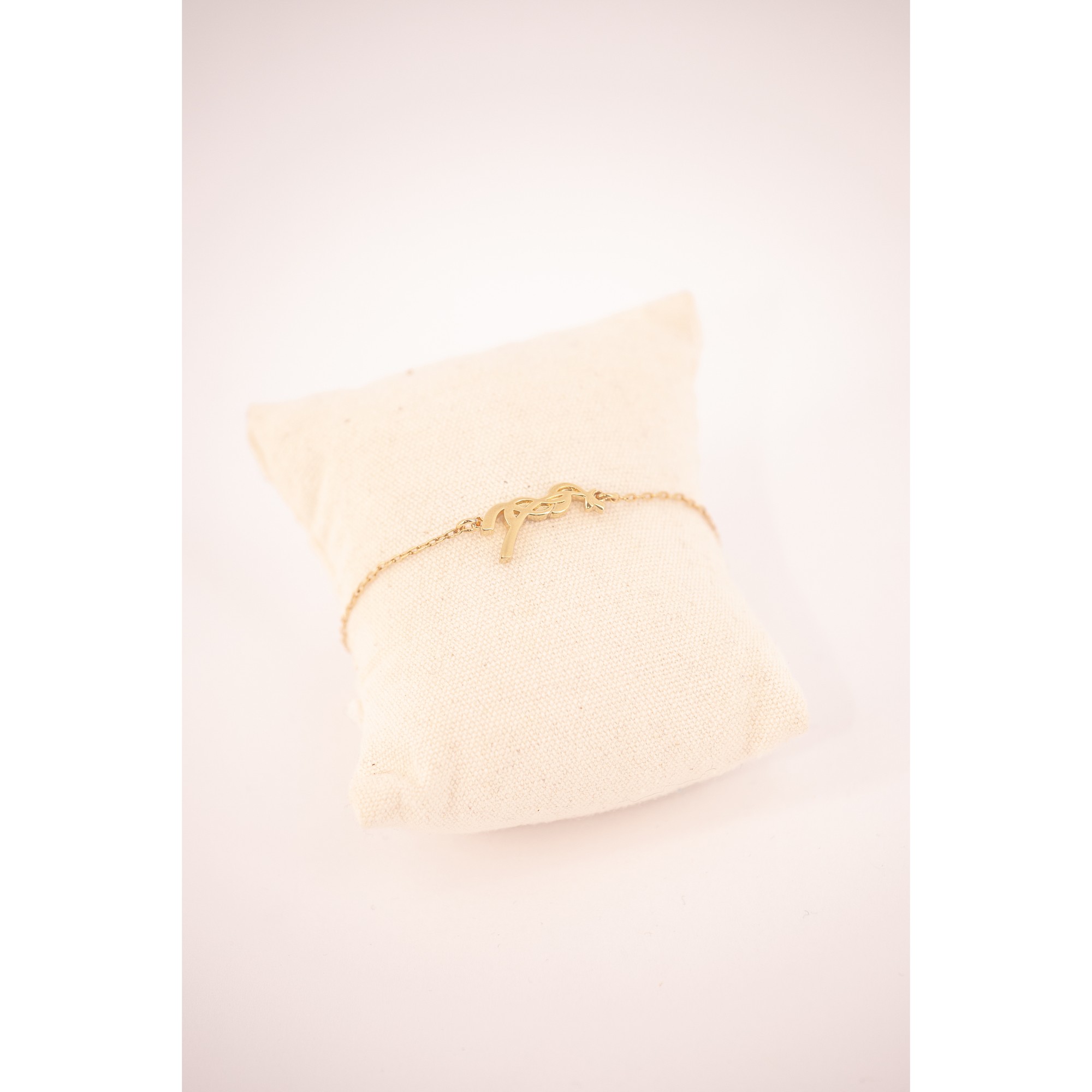 Gold bracelet with a delicate chain featuring a stylized letter "P," displayed on a soft, cream-colored cushion.