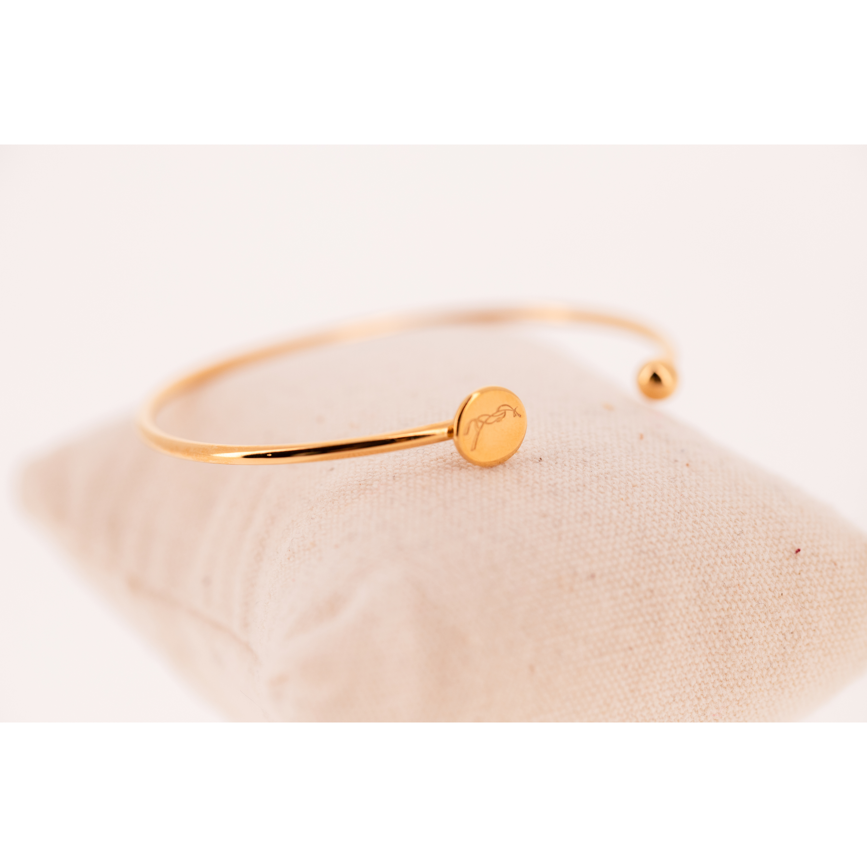 Gold-toned metal bracelet featuring a minimalist design with a small round charm and a smooth ball end, presented on a soft beige cushion.