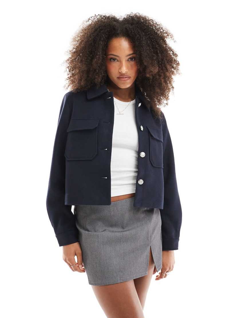 Navy blue cropped jacket with two front pockets, button closure, and a relaxed fit, styled over a white top and paired with a gray mini skirt.