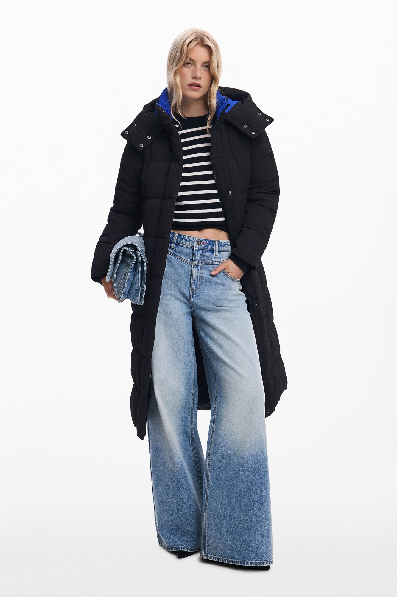 Long padded black coat with a belted waist, featuring a hood with blue lining, worn over a striped top and paired with wide-leg faded jeans.