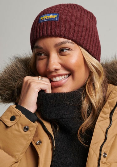 Maroon ribbed knit beanie with a blue and yellow Superdry logo patch, worn by a smiling woman in a tan jacket with a fur-lined hood.