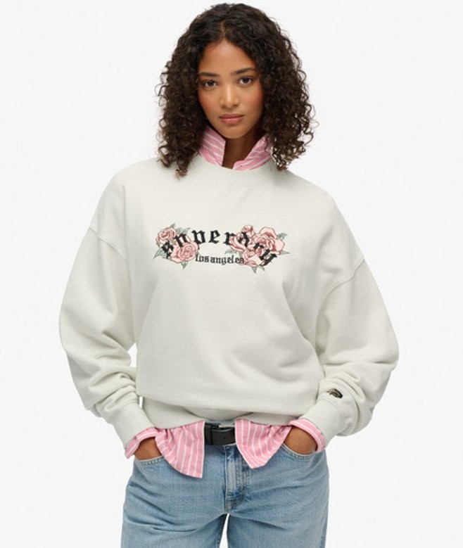 Cream oversized sweatshirt featuring floral "Superdry" logo, paired with a pink-striped collared shirt underneath, worn by a model with curly hair.