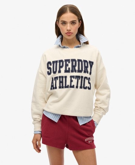 Cream-colored women's sweatshirt featuring "SUPERDRY ATHLETICS" in bold navy text, paired with a light blue collared shirt underneath and maroon shorts.