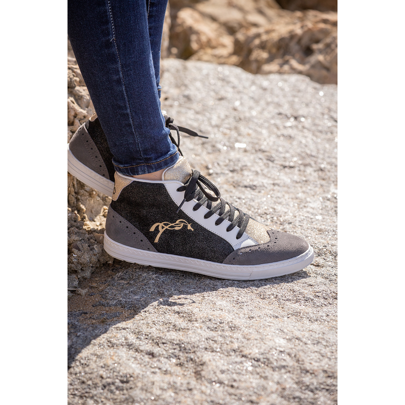 Stylish women's high-top trainers in black and grey with a textured finish, featuring a gold logo, white sole, and black laces, paired with dark denim jeans.