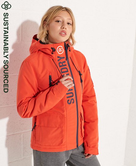 Bright orange waterproof jacket with a hood, featuring a prominent "SUPERDRY" logo along the front, zippered pockets, and a sustainable sourcing label.