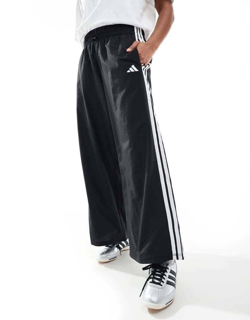 Black woven parachute trousers featuring three white stripes along the sides, an elastic waistband, and side pockets, styled with a relaxed fit.