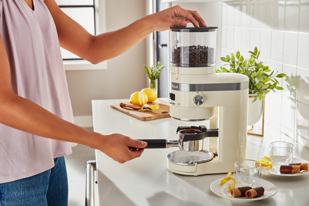 KitchenAid burr grinder in almond cream color, featuring a transparent hopper for beans, a metal portafilter holder, and a sleek design on a modern kitchen countertop.