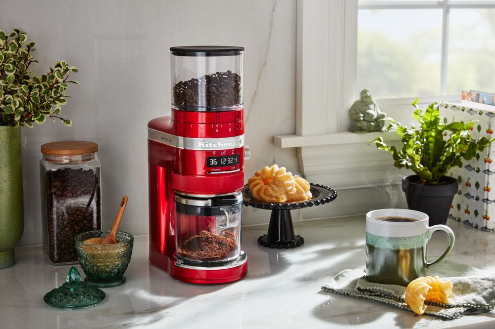 Candy apple red KitchenAid burr grinder features a transparent hopper, digital display, and a glass container for ground coffee, set on a marble countertop with coffee and pastries.