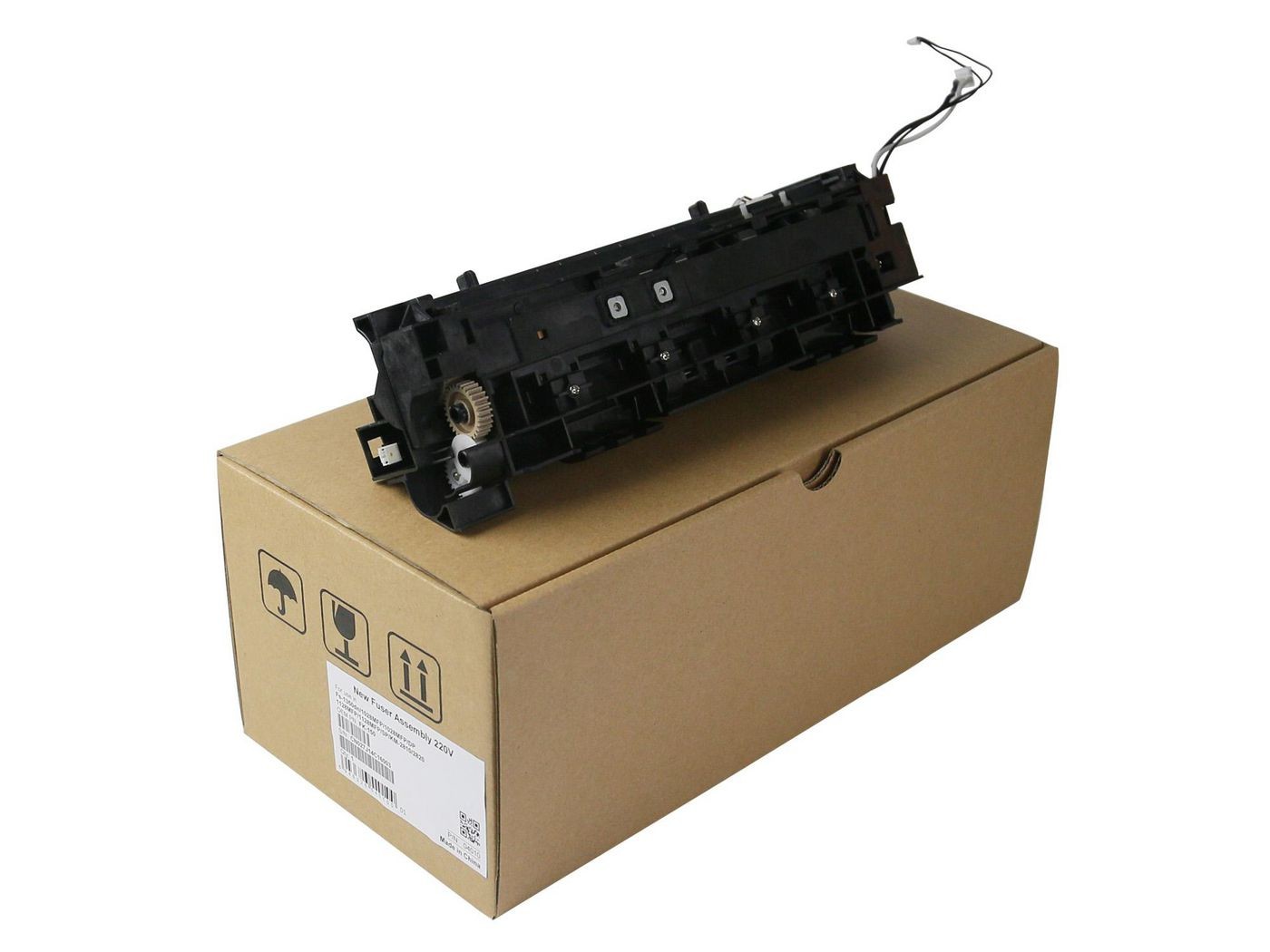 Black printer/scanner spare part with a wire, positioned on a brown cardboard box featuring handling icons and product information labels.
