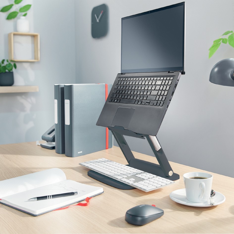 Height-adjustable Leitz Ergo Multi-Winkel Laptop Stand in gray, featuring a sleek design, supporting a laptop above a keyboard, with a notebook, pen, coffee cup, and mouse on a wooden desk.