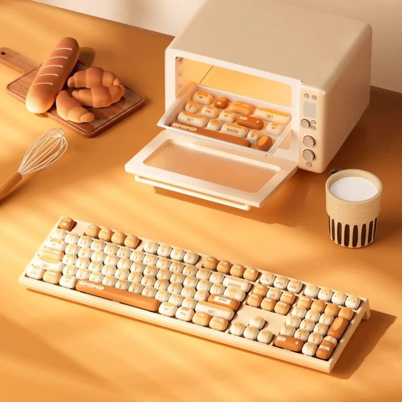 A beige and cream mechanical keyboard with cute pig-themed keycaps sits beside a miniature oven, surrounded by bread-shaped accessories and a cup.