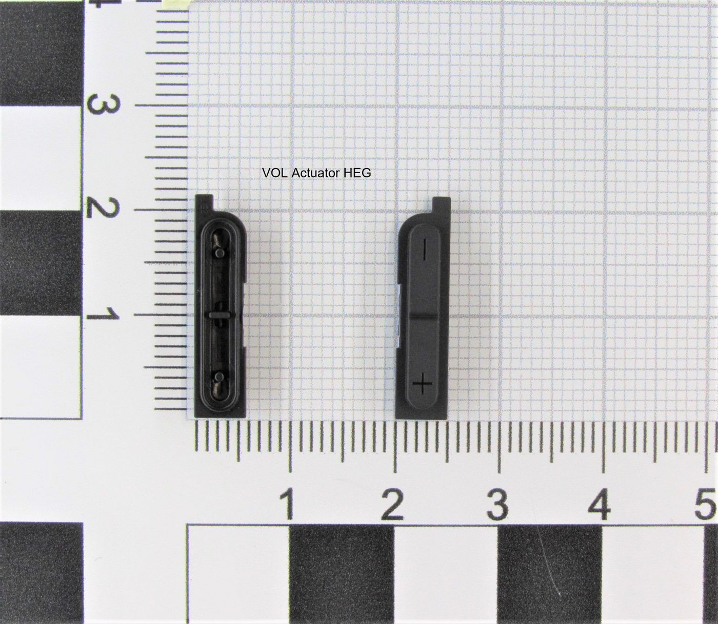 Black plastic volume actuator with two buttons, one marked with "+" and the other with a blank face, positioned on a grid background for scale.
