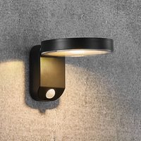 Round black Nordlux Rica LED solar wall light with sleek design and warm glow, mounted on a textured gray wall.