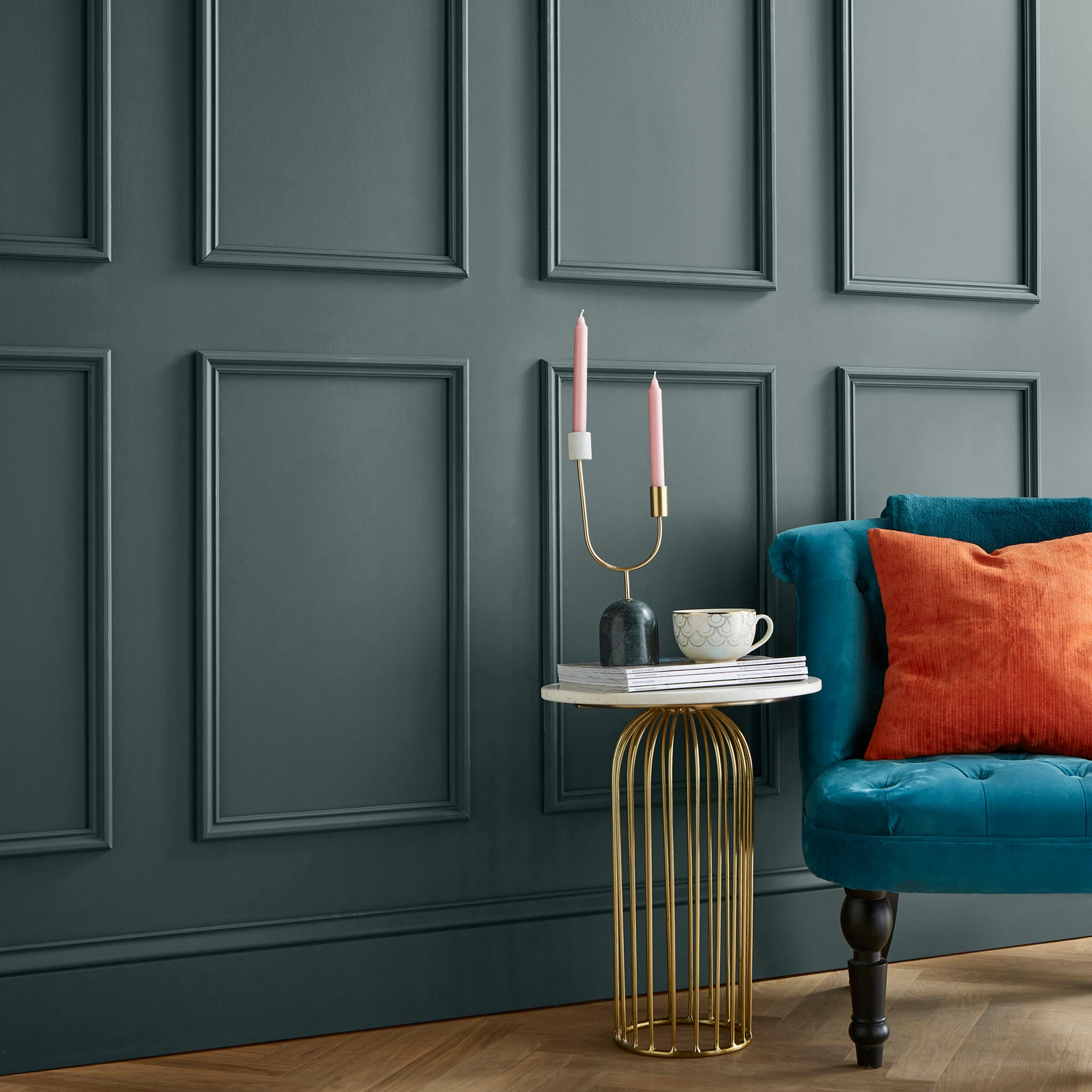 Painted dark green wall featuring a panel design, complemented by a teal velvet sofa with orange and patterned cushions, alongside a gold wire side table with candles and a cup.