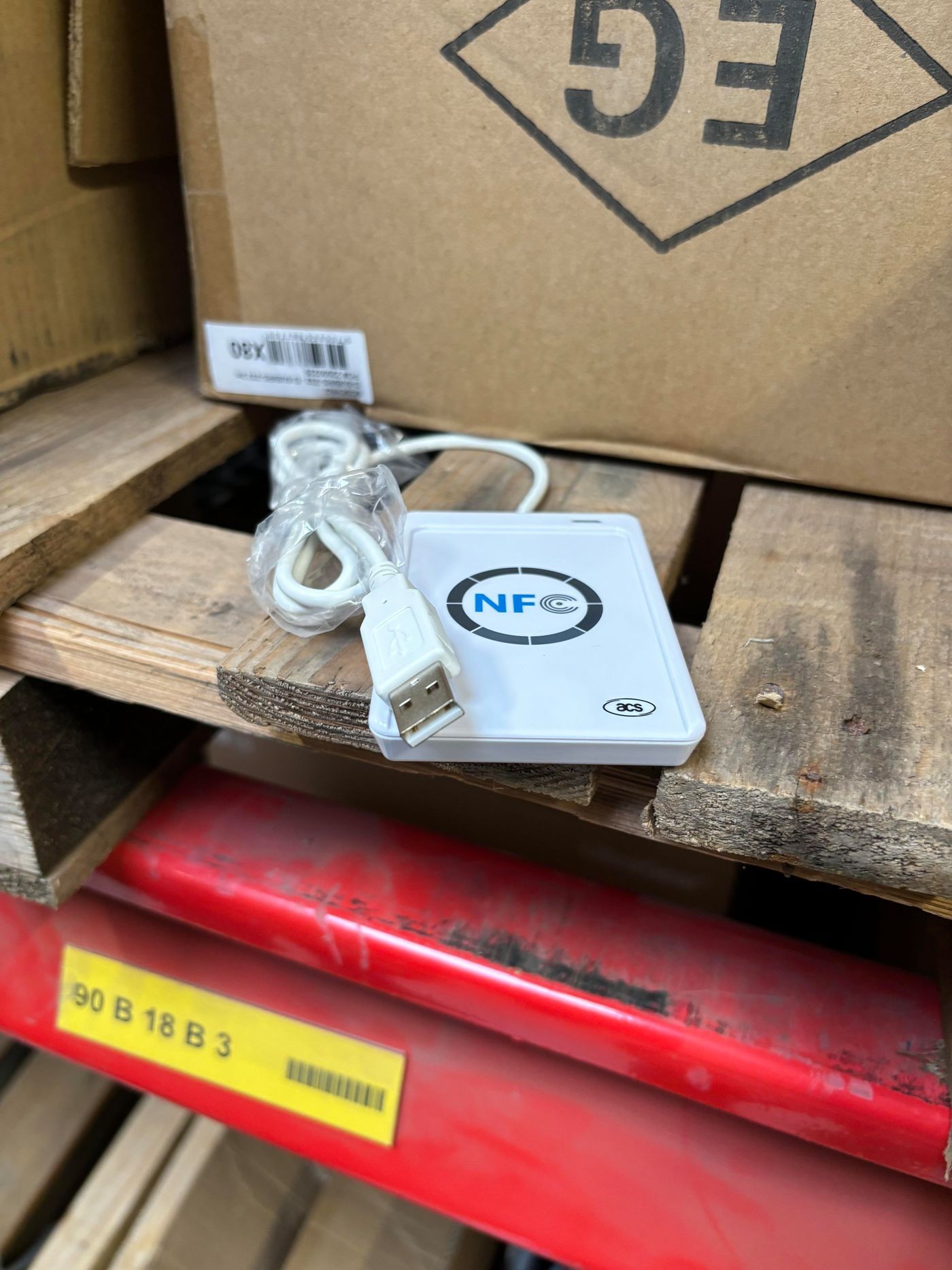 White NFC interface device with a USB connector, featuring a circular NFC logo on the top, resting on a wooden pallet beside cardboard boxes.