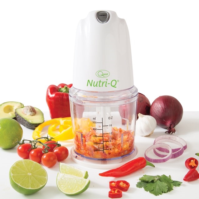White and clear plastic food chopper with a green "Nutri-Q" logo, featuring a measuring scale and surrounded by chopped vegetables like tomatoes, peppers, and onions.