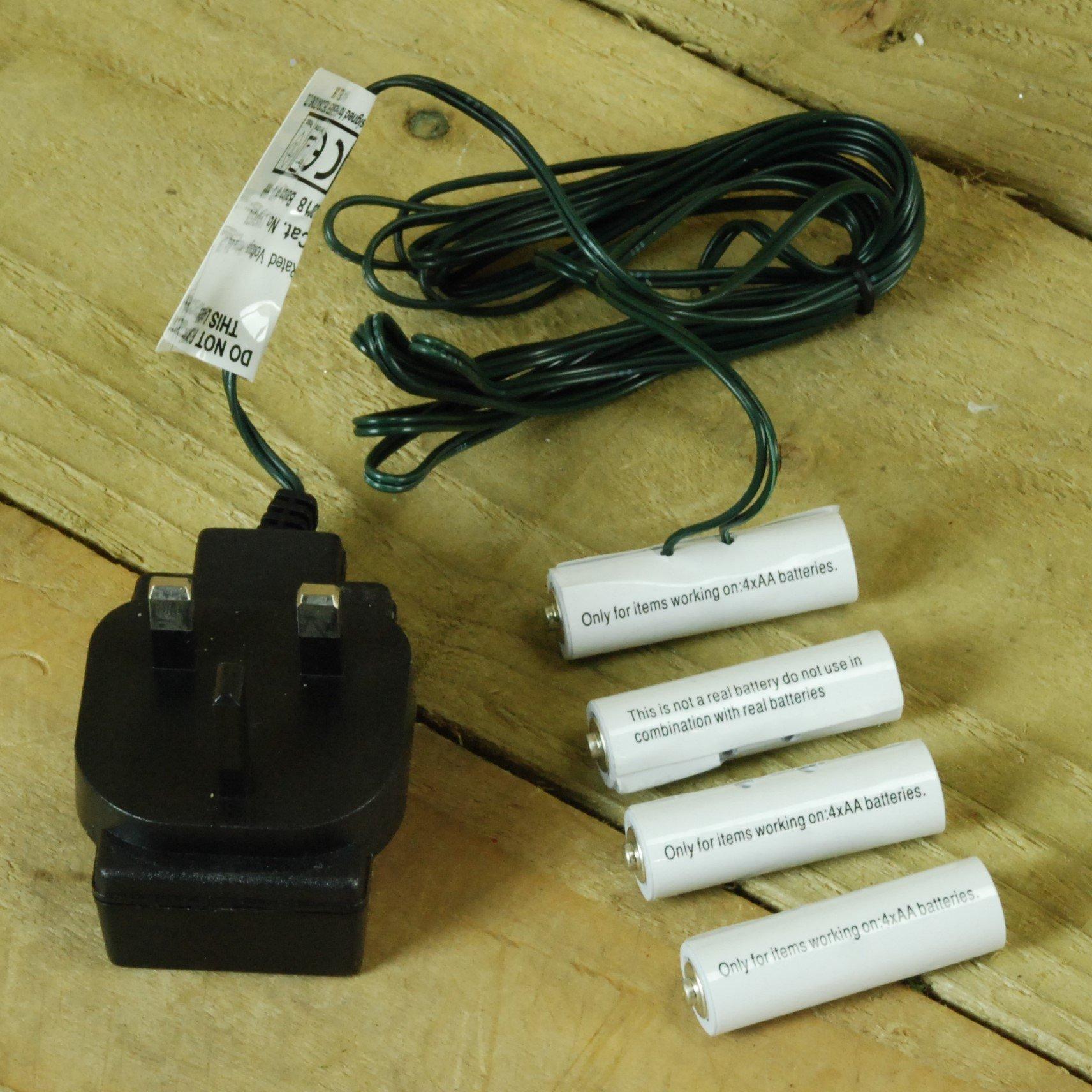 Black plug-in adapter with two prongs, accompanied by four cylindrical battery replacements and a green wire, labeled for compatibility with AA batteries.