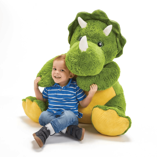 Green and yellow plush dinosaur, Digby, with soft fabric, large feet, and three white horns, sits beside a smiling child wearing a blue striped shirt and jeans.