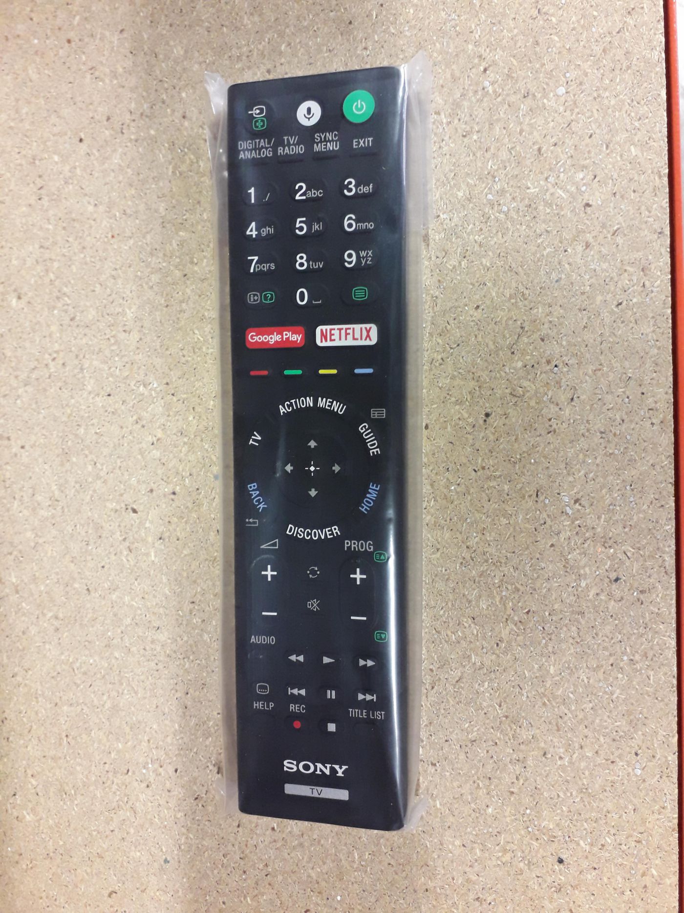 Black Sony remote control (RMF-X201ES) with a circular navigation pad, colorful buttons for Netflix and Google Play, and various function keys, wrapped in clear plastic.