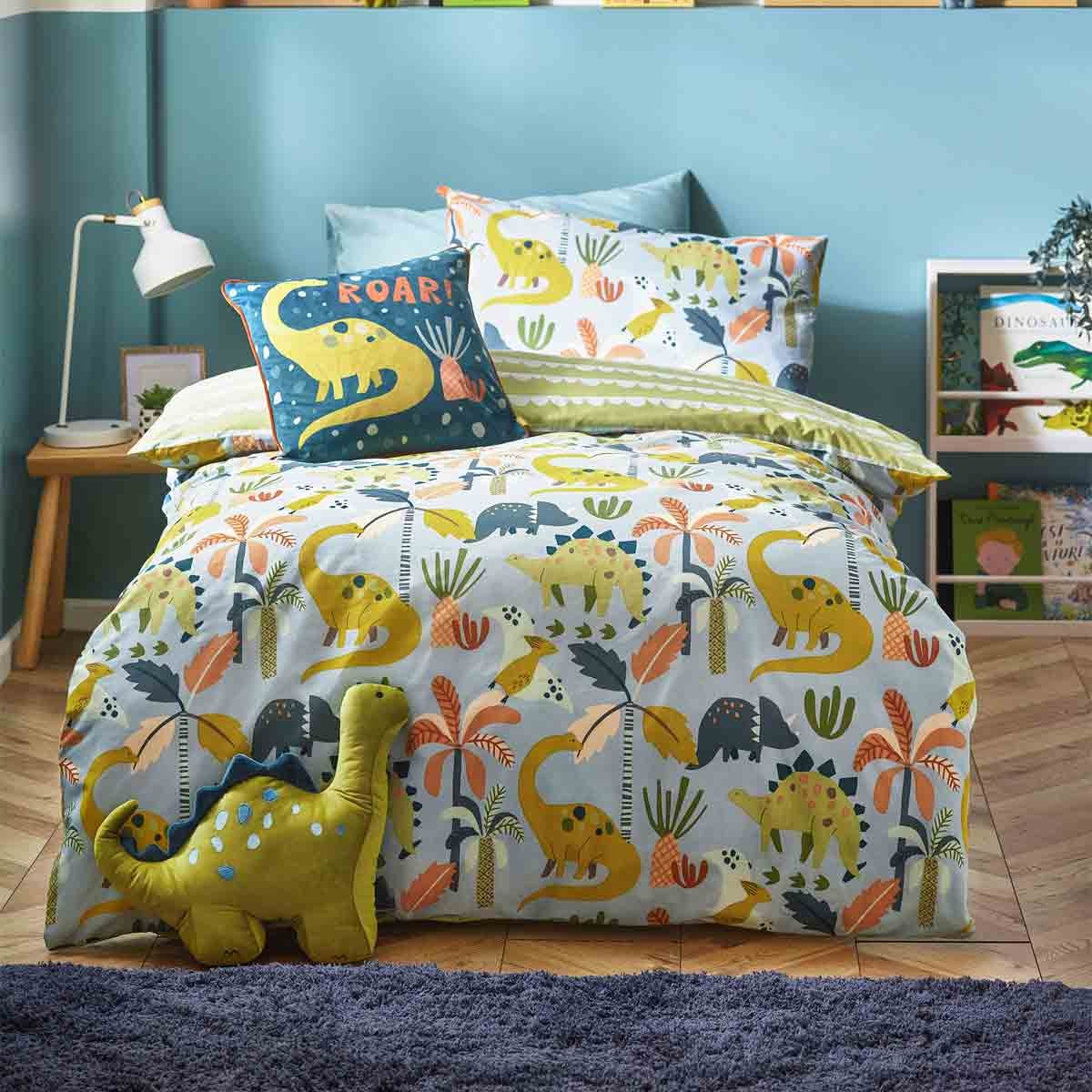 Scandi-style duvet set featuring playful blue and yellow dinosaur patterns on a soft fabric, complemented by a green plush dinosaur and a "ROAR!" cushion on a cozy bed.