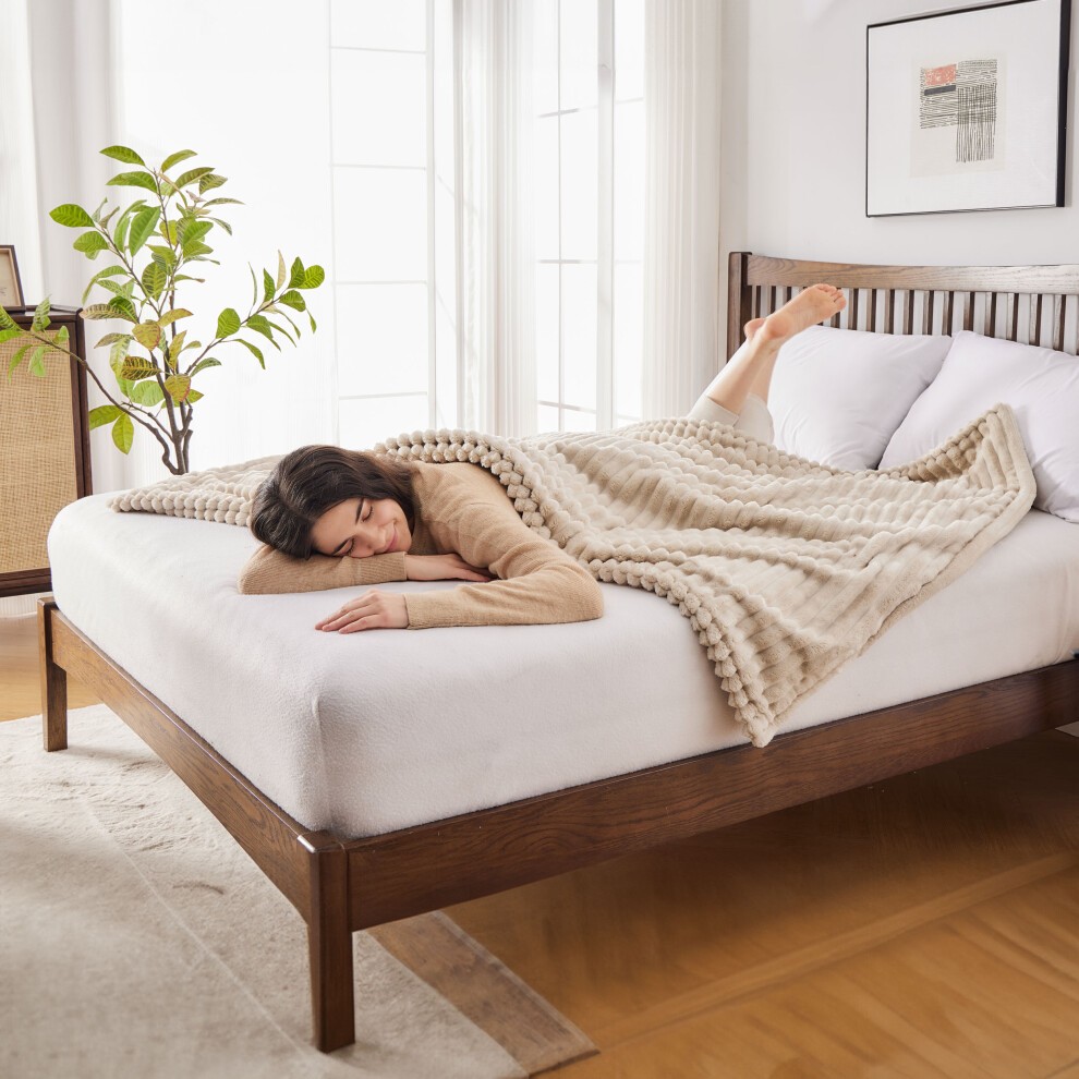 Cozy beige electric heated blanket with textured design draped over a bed, featuring a woman relaxing underneath, surrounded by natural light and indoor plants.
