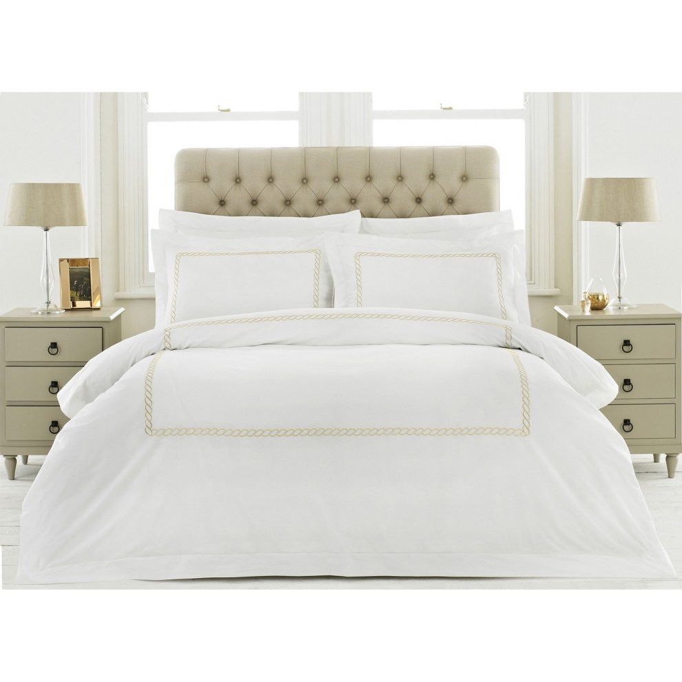 White cotton double duvet cover set with gold detailing, featuring a braided border, paired with two matching pillowcases, displayed on a neatly made bed with a beige tufted headboard.