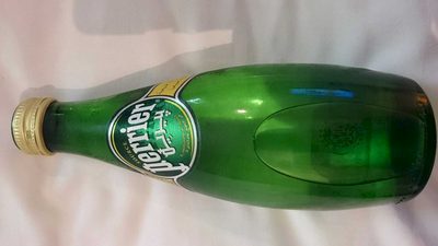 Green glass bottle of Perrier sparkling water, featuring a gold cap and a label with the brand name and logo, laid horizontally on a white surface.