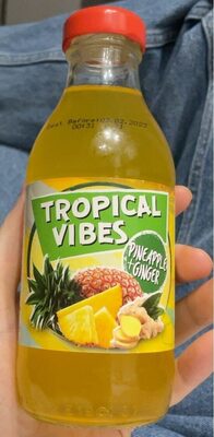 Clear glass bottle with a bright yellow drink, featuring a green label that reads "Tropical Vibes" with pineapple and ginger illustrations. Red cap on top.