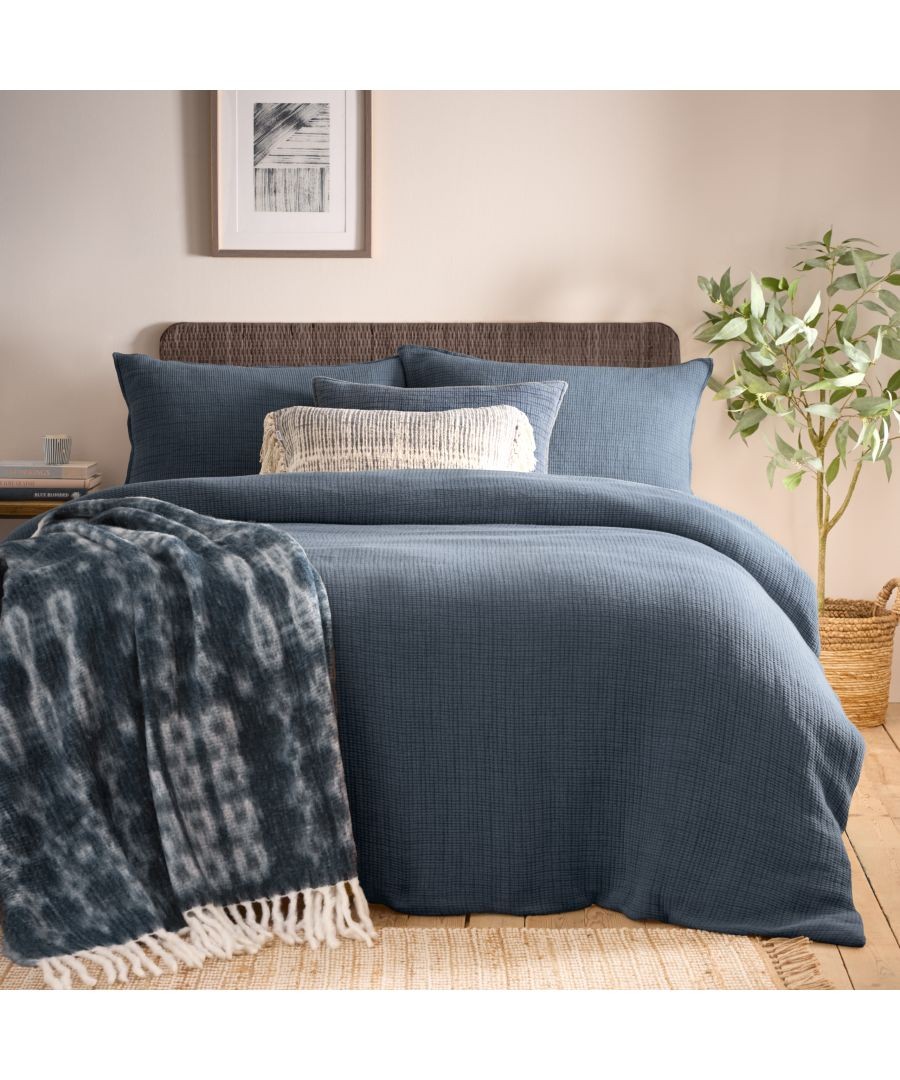 Blue acid-washed duvet set featuring a textured fabric, complemented by a decorative throw blanket with fringe, arranged on a bed with a natural wood headboard and greenery nearby.