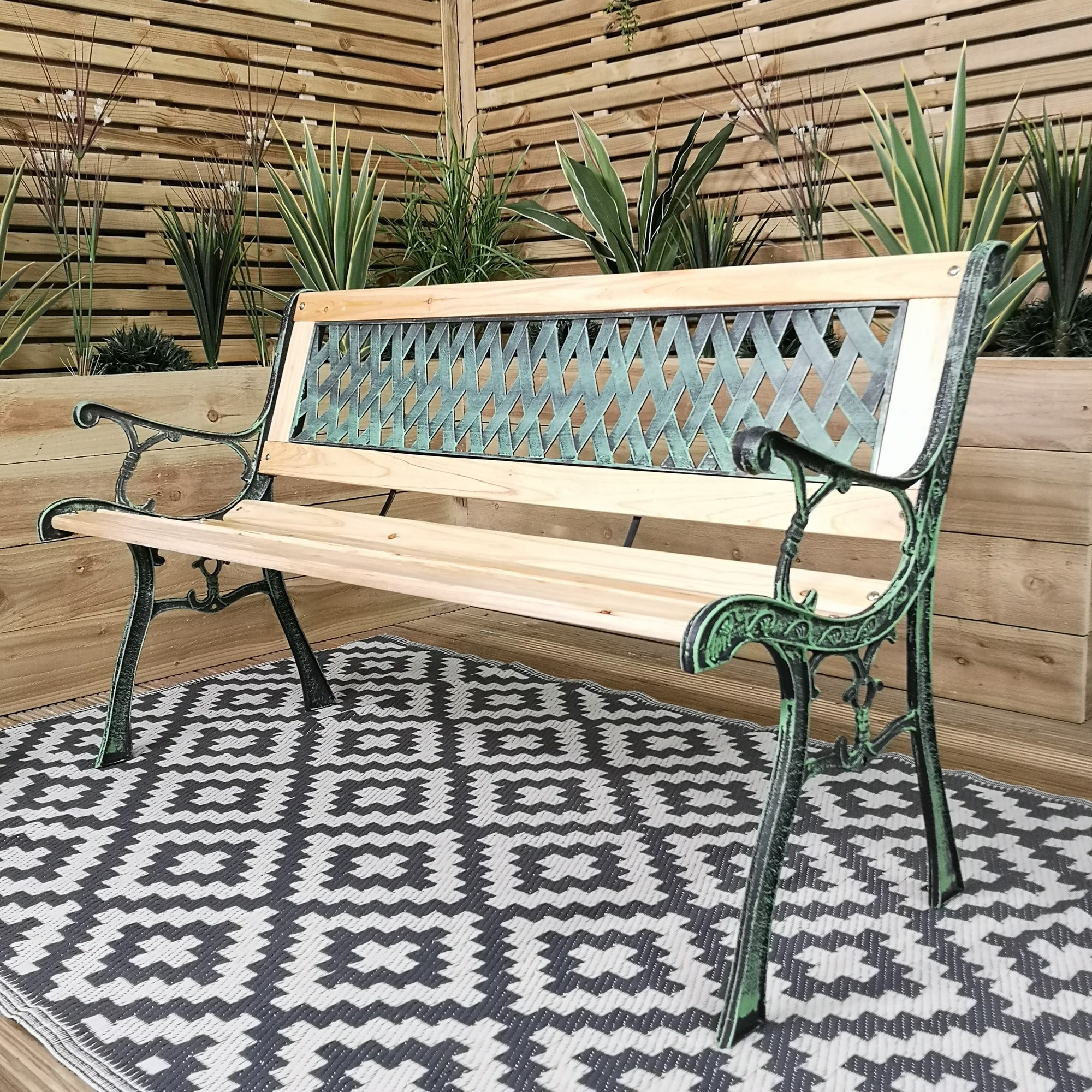 Brown wooden two-seater garden bench with a lattice design, featuring cast iron arms and legs, set against a backdrop of greenery and wooden paneling.