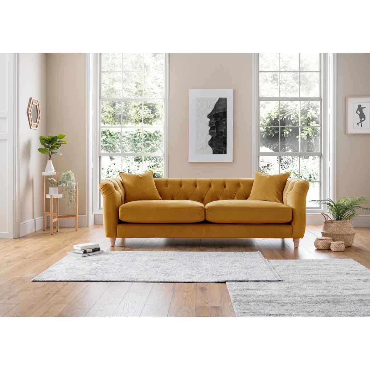 Large velvet sofa in ochre yellow with tufted back, two cushions, wooden legs, placed in a bright room with large windows and a light rug.